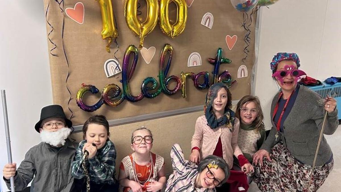 A group of children in old-timey constumes pose with their teacher who is also in an old-timey costume in front of a wall display of ballons and decorations that read, "100 Celebrate."