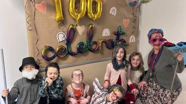 A group of children in old-timey constumes pose with their teacher who is also in an old-timey costume in front of a wall display of ballons and decorations that read, "100 Celebrate."