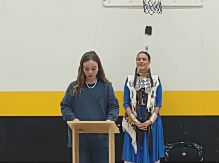A person standing at a podium with a person standing behind her
Description automatically generated
