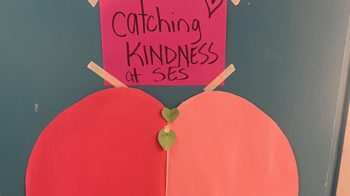 A two-tone red paper heart and pink label on a dark green background reading "Catching Kindness"