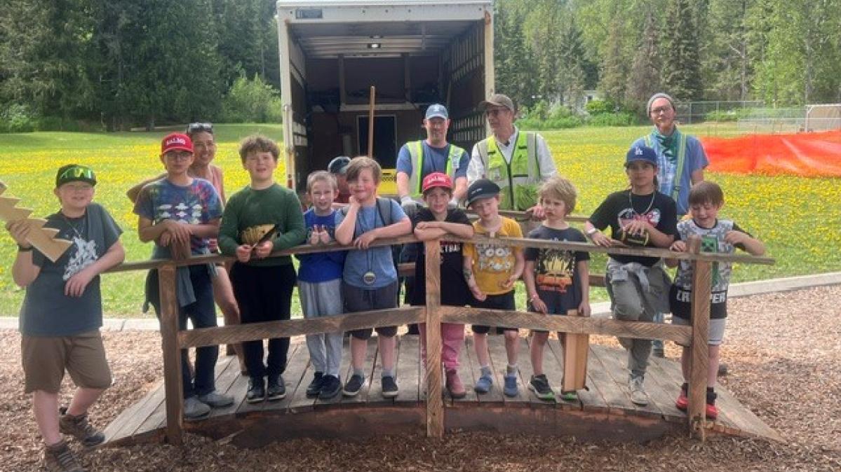 A group of elementary school students stand on a small wooden bridge with builders and teachers behind them