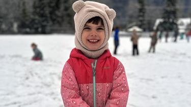 Primary school child in pink snowsuit and white hat with animal ears smiles in school yard on snowy day, with children playing in the background.