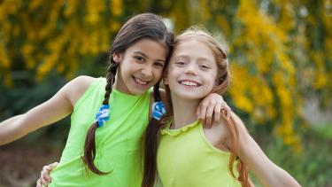 Two young girls wearing bright green and yellow sleeveless shirts and smiling broadly with arms around one anothers shoulders outdoors