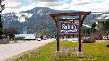 A wooden sign for small village on a green verge backed by a highway and snow covered mountains