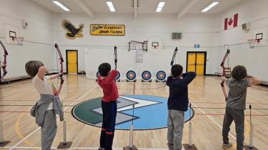 Four children in a row in a school gymnasium practicing archery