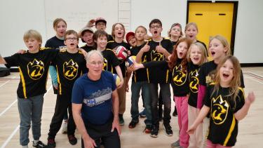 Elementary school students in matching black jersies cheering in a gymnasium with their teacher. 