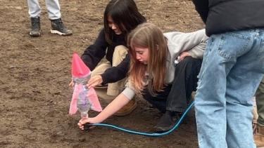 Elementary school students on a dirt field about to launch a small pink rocket