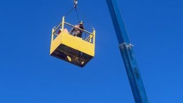 A crane holding a yellow rectangular cage holding a person high in the sky.
