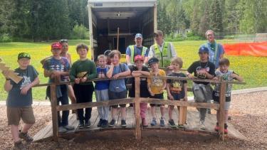 A group of elementary school students stand on a small wooden bridge with builders and teachers behind them