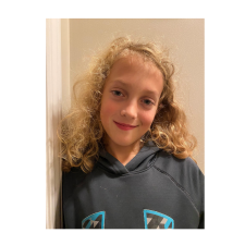 Girl with blonde, shoulder-length curly hair in navy sweatshirt leaning against a beige wall.