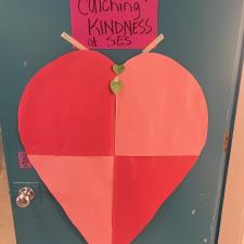 A two-tone red paper heart and pink label that reads, "Catching Kindness: against a green background