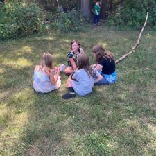 A group of children sit in a circle on the grass in front of a forest