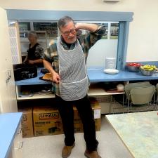 A man stands in a commercial kitchen wearing an apron and holding a flipper.