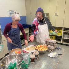 A woman in a blue apron and a man in a white apron chop fruit in a commercial kitchen.