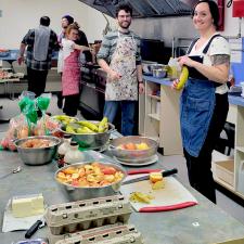 Five smiling people wearing aprons in a commercial kitchen cooking pancakes, preparing fruit with a bowl of apples, butter and cartons of eggs in the foreground on a table.