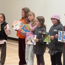 Elementary school students stand in a gymnasium holding up artwork on paper for others to see