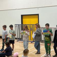 Elementary school students stand in a gymnasium holding up artwork for others to see