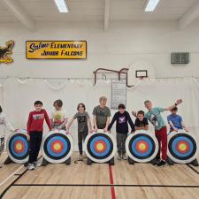 Elementary school students stand in a gymnasium behind archery targets that show evidence of their archery abilities.