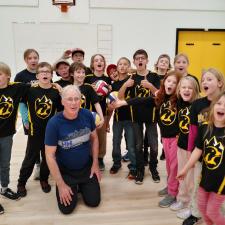 Elementary school students in matching black jersies cheering in a gymnasium with their teacher. 