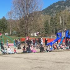 A group of children and adults sitting in a dirt playground with trees and mountains in the background in spring