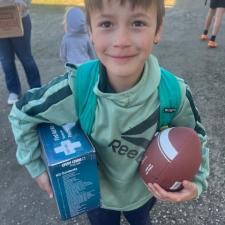 A boy in jeans and green hoodie holds a football in one hand and a blue box under one arm.
