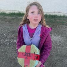 A girl in a magenta jacket and mauve sweater holds a yellow and red taped-up box outdoors in front of a white wall.