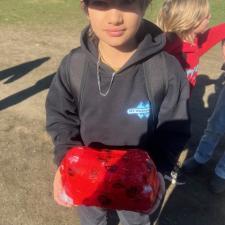 A boy in a blue ball cap and black hoodie holds a red package outdoors in a school field