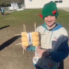 A boy in a green toque with red horns and wearing a beige and blue hoody holds a square foam contraption with wooden skewers sticking out of it.