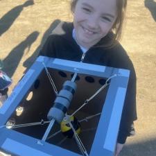 A girl in a black hoody holds a blue contraption with guy wires and other donut-type features, outside in the sunshine.