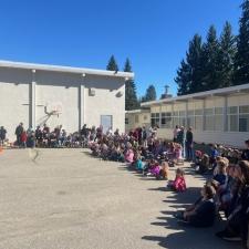 Elementary school students seated in a semi-circle in a paved parking lot in front of an elementary school gym and classrooms