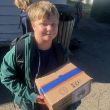A boy in a green jacket and black backpack holds a cardboard box outdoors in the sunshine in front of a school building