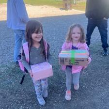 Two girls in pink, grey and blue clothing hold cardboard boxes in a school parking lot.