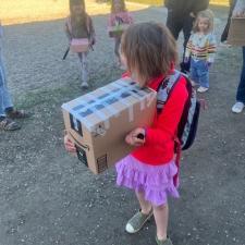A girl in a pink skirt and red jacket holds a big cardboard box outdoors in a parking lot with children and adults in the background.