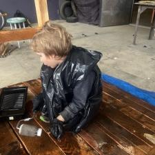 A blonde elementary student wearing a garbage bag stains a wooden bridge sitting on a tarp in a school area outdoors