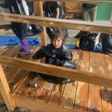 A brown haired elementary student wearing a garbage bag stains a wooden bridge sitting on a tarp in a school area outdoors