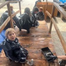 A group of elementary school students wearing garbage bags stain a wooden bridge sitting on a tarp in a school area outdoors