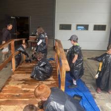 A group of elementary school students wearing garbage bags stain a wooden bridge sitting on a tarp in a school area outdoors