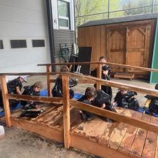 A group of elementary school students wearing garbage bags stain a wooden bridge sitting on a tarp in a school area outdoors