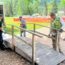 Men in safety vests carry a wooden bridge structure into a school yard