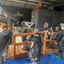 A group of elementary school students wearing garbage bags stain a wooden bridge sitting on a tarp in a school area outdoors