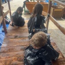 A group of elementary school students wearing garbage bags stain a wooden bridge sitting on a tarp in a school area outdoors