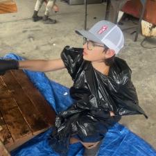 An elementary student wearing a garbage bag and trucker cap stains a wooden bridge sitting on a tarp in a school area outdoors